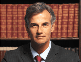 Man in a suit with gray hair stands in front of a row of books on a shelf.