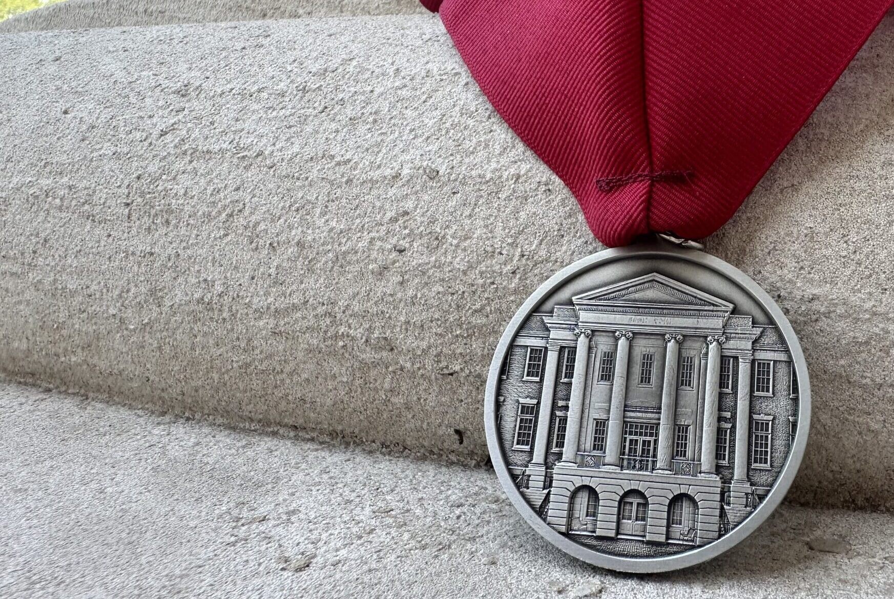 A silver medal featuring an image of a neoclassical building and the name Gordon J. Glover hangs from a maroon ribbon, resting on stone steps.