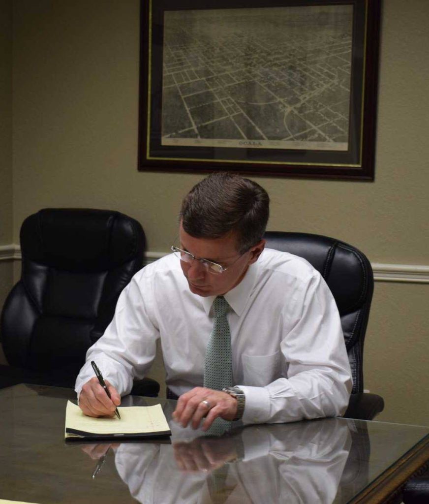 A man in a white shirt and tie sits at a desk writing on a notepad, with an empty chair beside him and a framed map on the wall behind—reminiscent of Gordon J. Glover’s composed office moments.